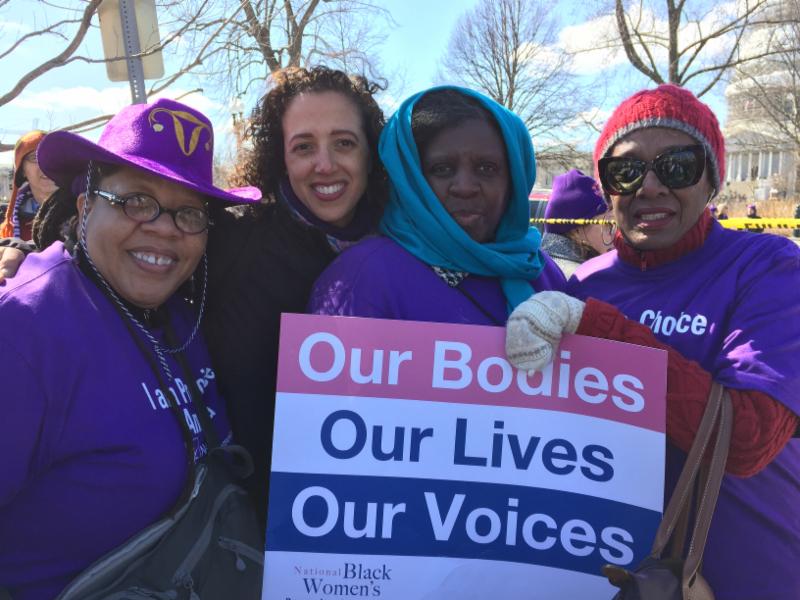 Photo of women gathered outside holding a sign reading "Our Bodies, Our Lives, our Voices."